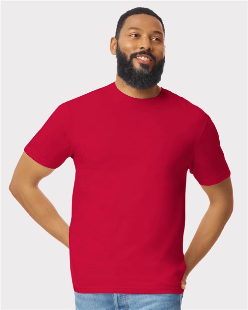 A bearded man is smiling as he stands against a plain white backdrop, hands on hips, in a plain red Gildan - Softstyle® T-Shirt made of ring-spun cotton and light blue jeans.