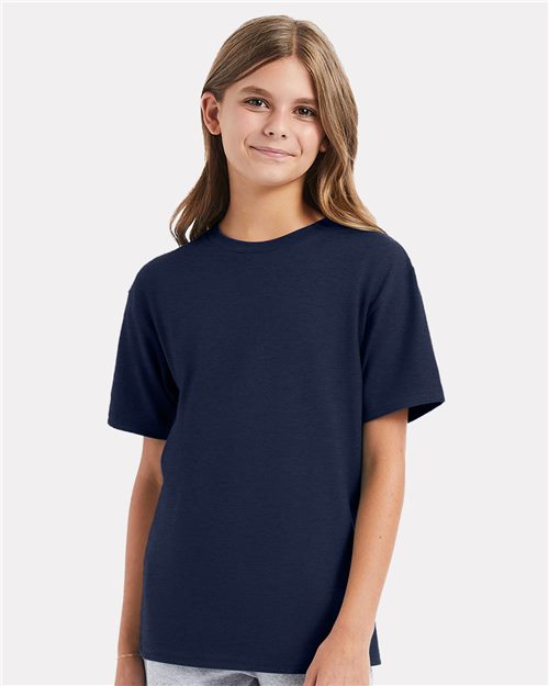 Load image into Gallery viewer, A young person with long hair stands against a light gray background, wearing the navy blue Hanes - Perfect-T Youth T-Shirt made of ring-spun cotton and light blue jeans, smiling slightly at the camera.
