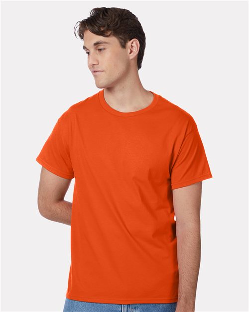 A person wearing a Hanes - Authentic T-Shirt, dyed orange, along with blue jeans stands against a plain white background. Looking off to the side with one arm behind their back, they capture the spirit of socially conscious manufacturing.