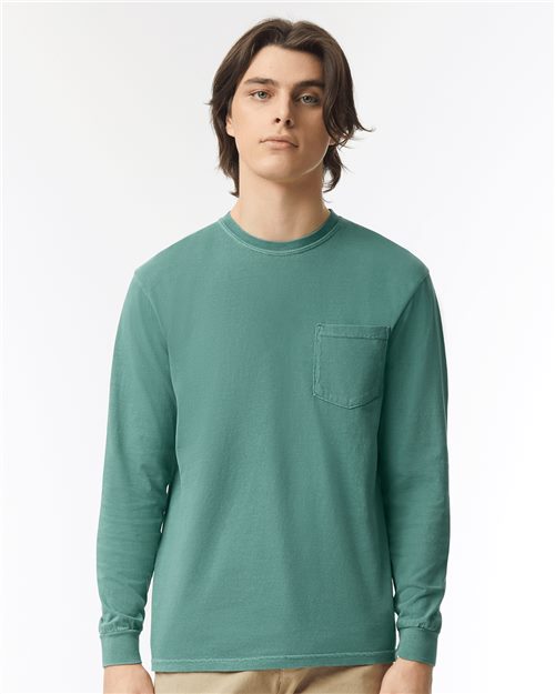 A person with long brown hair is wearing a teal Comfort Colors Garment-Dyed Heavyweight Long Sleeve Pocket T-Shirt made from ring-spun cotton. The shirt has a pocket on the left side. The background is plain white, and the individual is looking directly at the camera.