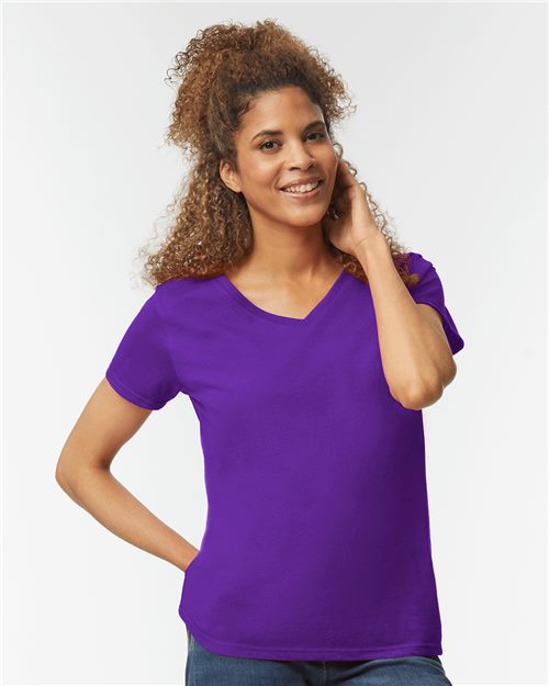 A person with curly hair smiles in a Gildan Heavy Cotton Women’s V-Neck T-Shirt made from U.S. cotton, and jeans. The plain white background highlights the sustainable production of this bright purple garment.