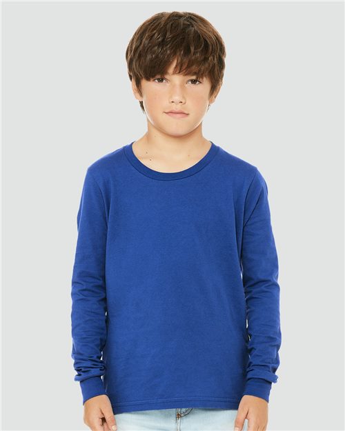 Load image into Gallery viewer, A young boy with brown hair stands against a light gray background, wearing a BELLA + CANVAS Youth Jersey Long Sleeve Tee in blue, made from Airlume combed cotton, paired with light blue jeans. He has a neutral expression, hands by his sides, showcasing the unisex sizing and sustainable manufacturing of the brand.
