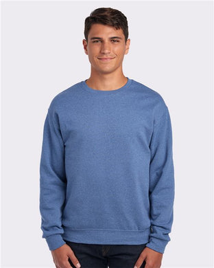 A person in a blue JERZEES - NuBlend® Crewneck Sweatshirt and dark jeans stands against a plain white background, smiling at the camera.