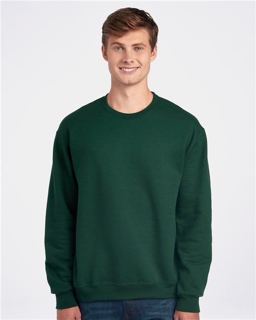 A young person wears a JERZEES - Super Sweats NuBlend® Crewneck Sweatshirt, a pill-resistant fleece in dark green. They stand against a light gray background, smiling with short brown hair and dressed in blue jeans.