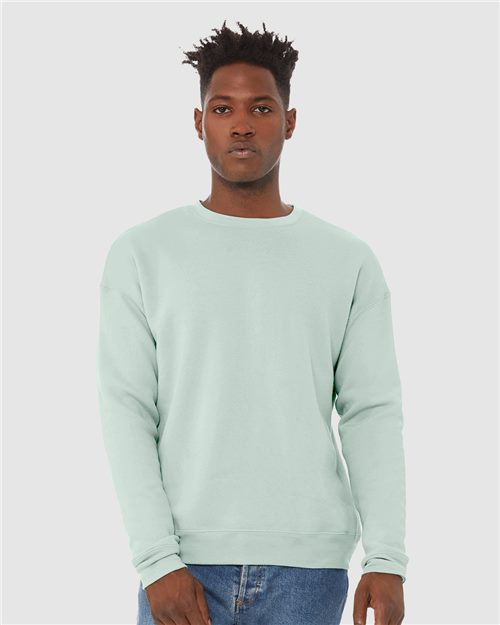 Load image into Gallery viewer, Wearing a light mint green BELLA + CANVAS Sponge Fleece Drop Shoulder Crewneck Sweatshirt made from Airlume combed and ring-spun cotton, a person pairs it with blue jeans against a plain gray background, exuding confidence and embodying sustainable manufacturing while looking directly at the camera.
