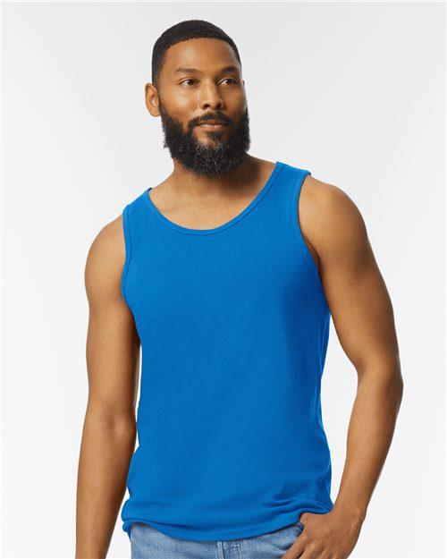 A man with a beard and short hair wears a bright blue Gildan - Heavy Cotton™ Tank Top and light blue jeans. He stands against a plain white background, looking slightly to the side and exuding an effortlessly cool vibe.