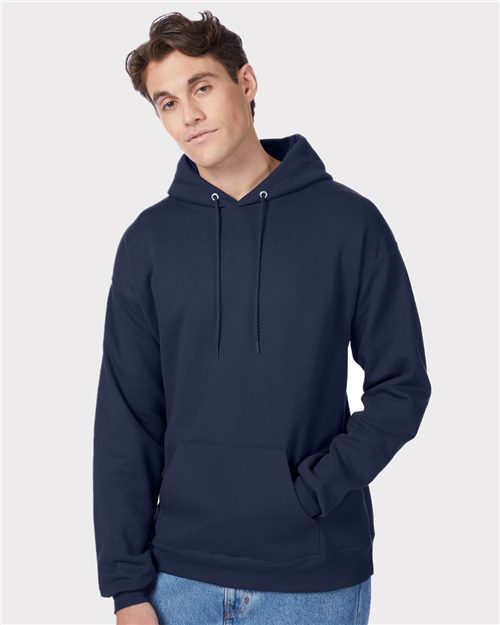 Someone stands against a white background wearing a Hanes Ecosmart® Hooded Sweatshirt made from a sustainable cotton/polyester blend. With the hood down and one hand in their pocket, they pair it effortlessly with light blue jeans and wear a relaxed expression.