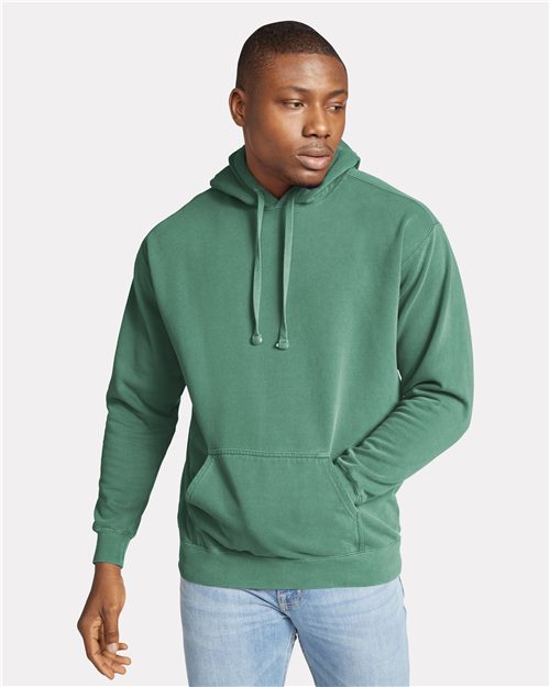 Load image into Gallery viewer, A person is wearing a Comfort Colors Garment-Dyed Hooded Sweatshirt, crafted from soft cotton face fleece, with the hood down and light blue jeans. They have one hand in the pocket and are looking slightly to the side against a plain gray background.
