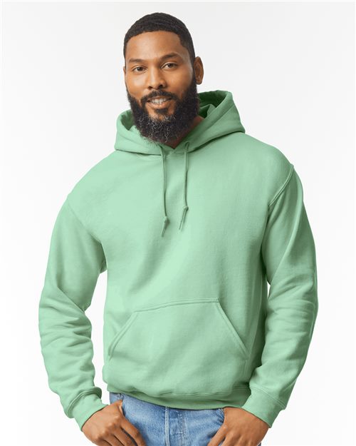 The bearded individual, wearing a light green Gildan Heavy Blend™ Hooded Sweatshirt and jeans, stands against a plain white background. With hands in pockets and smiling confidently at the camera.