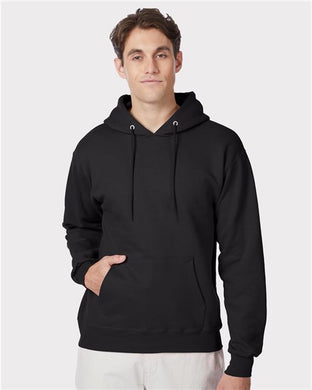 A person wears a Hanes Ultimate Cotton® Hooded Sweatshirt, made from a cotton/polyester blend with sustainable practices, against a light gray background. The black hoodie features drawstrings and a front pocket, paired stylishly with light-colored pants.