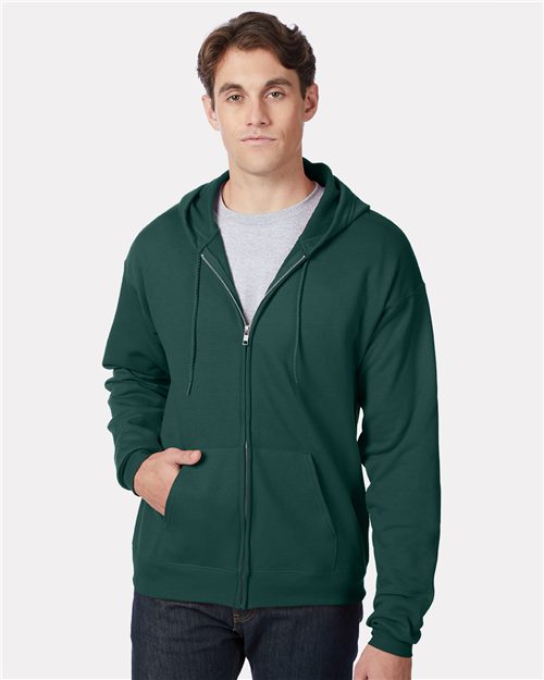 Load image into Gallery viewer, A person in a dark green Hanes - Ecosmart® Full-Zip Hooded Sweatshirt, a light gray T-shirt, and dark jeans stands against a plain background with one hand in their pocket and a neutral expression.
