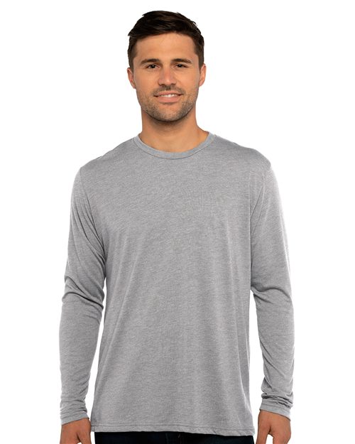 A person with short dark hair wears a Next Level - Triblend Long Sleeve T-Shirt, showcasing socially conscious manufacturing. They're standing and smiling against a plain white background, wearing dark pants. The shirt is crafted from polyester, combed ring-spun cotton, and rayon.