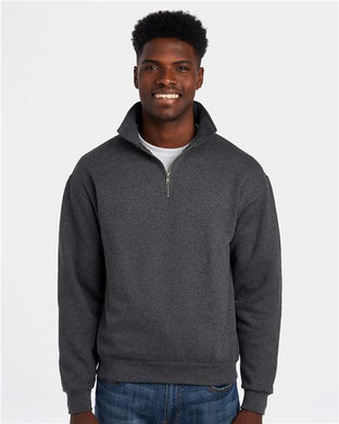 A smiling person wearing a JERZEES - Super Sweats NuBlend® Quarter-Zip Cadet Collar Sweatshirt in dark gray stands against a white background. The sweater features socially conscious manufacturing, pill-resistant fleece, and a high collar while they rest their hands by their sides over a white shirt and blue jeans.