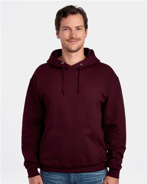 A person wears a JERZEES Super Sweats NuBlend® Hooded Sweatshirt and blue jeans, standing with a smile and hands slightly in pockets, set against a simple white background.