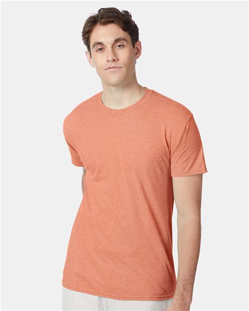 A person in an orange Hanes Perfect-T Triblend T-Shirt stands against a white background, paired with light-colored pants. They sport short dark hair and a neutral expression, and their outfit showcases a modern fit. The t-shirt is made from recycled polyester.