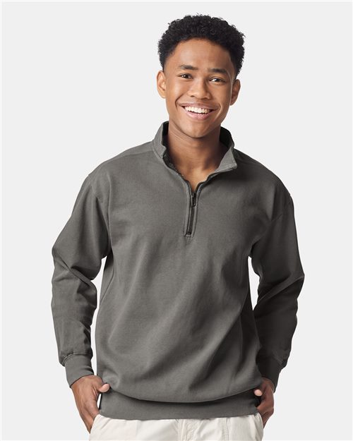 A person smiles while wearing a Comfort Colors dark gray garment-dyed quarter-zip sweatshirt and light-colored pants, standing against a plain background.