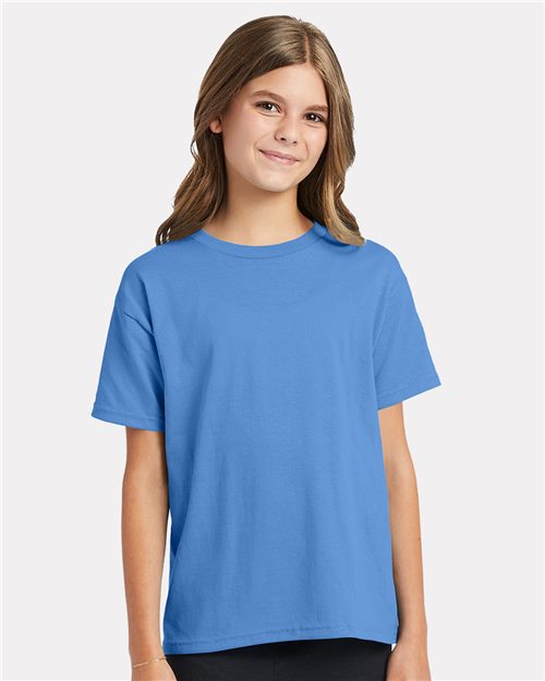 A young person with long brown hair, wearing a Hanes Ecosmart™ Youth T-Shirt in plain blue and black pants, smiles against a white background.