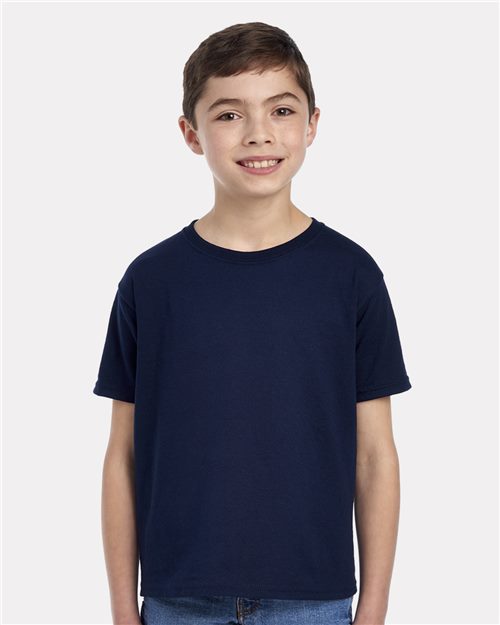 A young boy with short brown hair is smiling at the camera, wearing a JERZEES - Dri-Power® Youth 50/50 T-Shirt and blue jeans, standing against a plain white background.