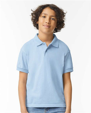 A young person with curly hair stands against a plain white background, wearing a Gildan - DryBlend® Youth Jersey Polo in light blue and jeans, smiling gently.