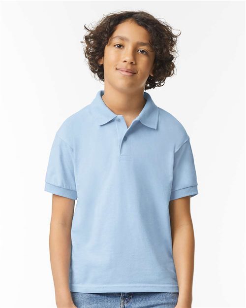 Load image into Gallery viewer, A young person with curly hair stands against a plain white background, wearing a Gildan - DryBlend® Youth Jersey Polo in light blue and jeans, smiling gently.
