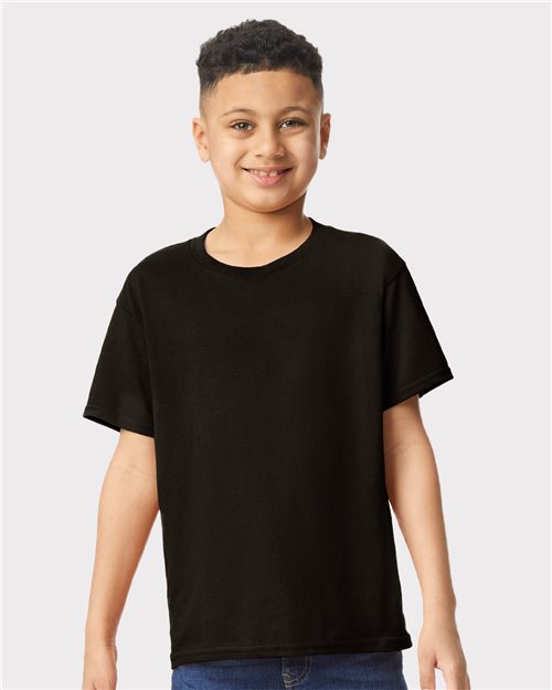 A young person beams in a Gildan - Heavy Cotton™ Youth T-Shirt and blue jeans. Their short, curly hair adds charm as they stand against a light gray backdrop, exuding comfort and style thanks to sustainable manufacturing practices.