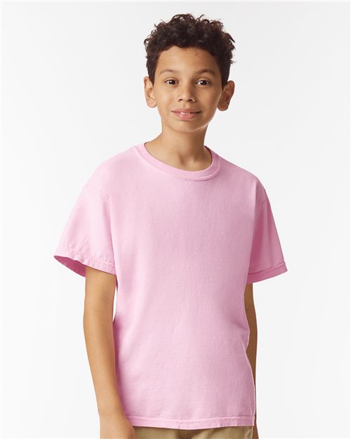 A young boy with curly hair wears a pink Comfort Colors - Garment-Dyed Youth Heavyweight T-Shirt and beige pants, standing against a white background while smiling softly.