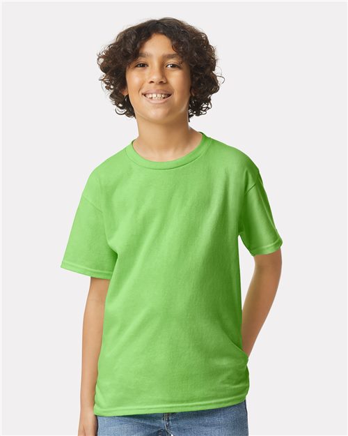 A smiling child with curly hair, wearing a bright green Gildan Ultra Cotton Youth T-Shirt and blue jeans, stands against a plain background, looking at the camera.