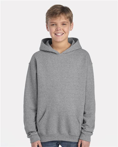A young boy wearing a JERZEES - NuBlend® Youth Hooded Sweatshirt smiles at the camera with hands casually in the pouch pocket. Made from pill-resistant fleece, this gray hoodie is set against a plain light gray background and is produced through sustainable practices.