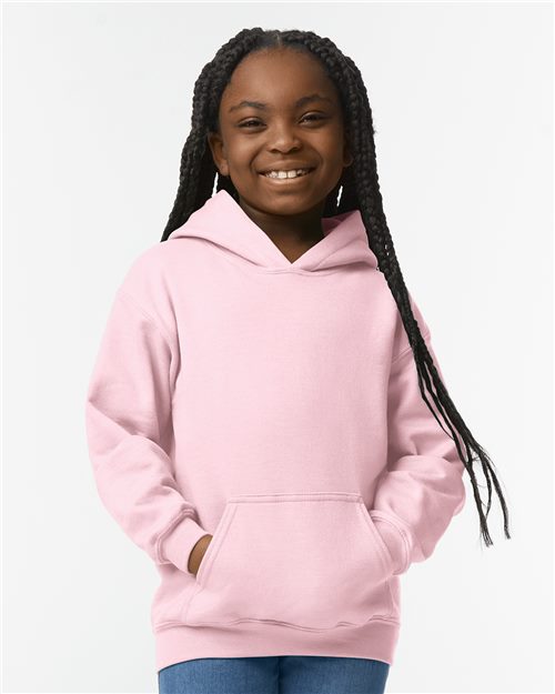 A girl with long braided hair wears a pink Gildan - Heavy Blend™ Youth Hooded Sweatshirt and blue jeans, standing against a white background, embodying sustainable manufacturing.