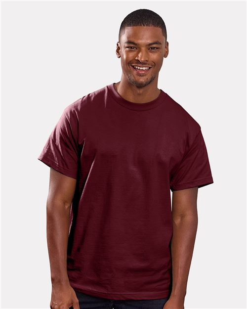 A smiling person in a Hanes Beefy-T® T-shirt, crafted from heavyweight cotton, stands against a light gray backdrop, highlighting its stylish and sustainable design.