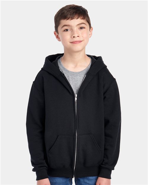 A young boy wears a JERZEES NuBlend® Youth Full-Zip Hooded Sweatshirt made of sustainably sourced cotton with high-stitch density. He stands against a plain light gray backdrop, gazing neutrally at the camera, finishing his outfit with a gray t-shirt and blue jeans.