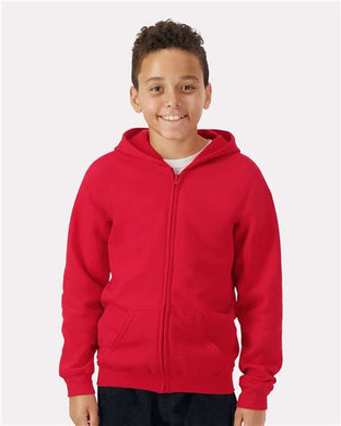 A smiling child in a Gildan - Heavy Blend™ Youth Full-Zip Hooded Sweatshirt and dark pants stands against a plain white backdrop, highlighting sustainable manufacturing.