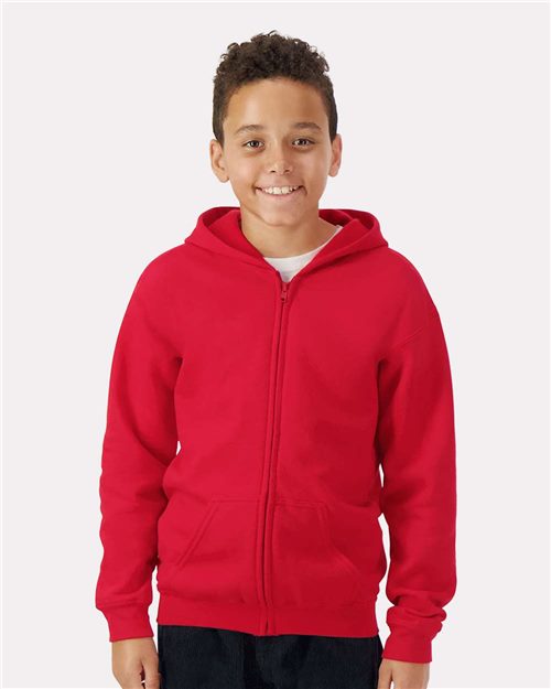 Load image into Gallery viewer, A smiling child in a Gildan - Heavy Blend™ Youth Full-Zip Hooded Sweatshirt and dark pants stands against a plain white backdrop, highlighting sustainable manufacturing.
