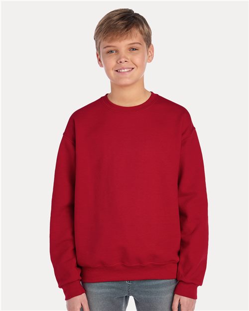 Load image into Gallery viewer, A young boy with short hair smiles while wearing a JERZEES - NuBlend® Youth Crewneck Sweatshirt in plain red, crafted from pill-resistant fleece, along with jeans. He stands against a white background, showcasing sustainable manufacturing through his eco-friendly attire.
