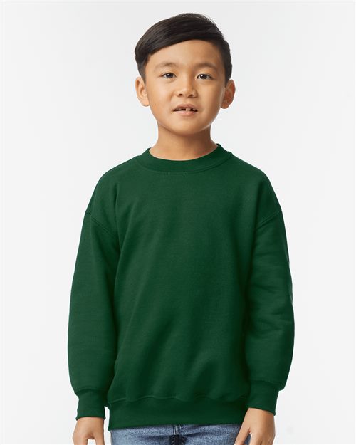 A young boy in a Gildan Heavy Blend™ Youth Sweatshirt, dark green, paired with blue jeans stands against a plain white background. With short dark hair and a neutral expression, he embodies the spirit of sustainable manufacturing.