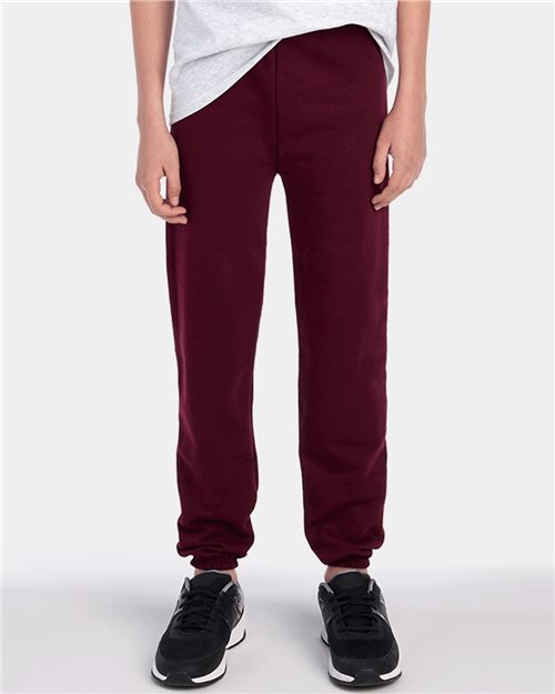 A person wearing JERZEES - NuBlend® Youth Sweatpants in burgundy, made from pill-resistant fleece, stands against a light gray background with black sneakers. Their hands rest at their sides, complemented by a partially visible light gray top.