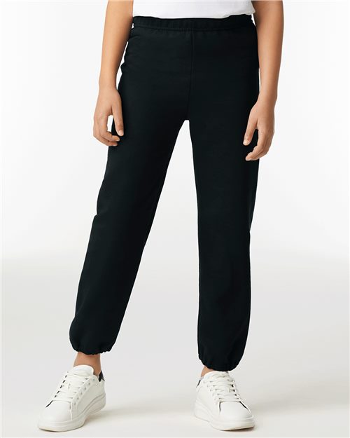 A person wearing black Gildan - Heavy Blend™ Youth Sweatpants with an elastic waistband and cuffs, paired with white sneakers, stands against a plain white background, highlighting comfort and style.