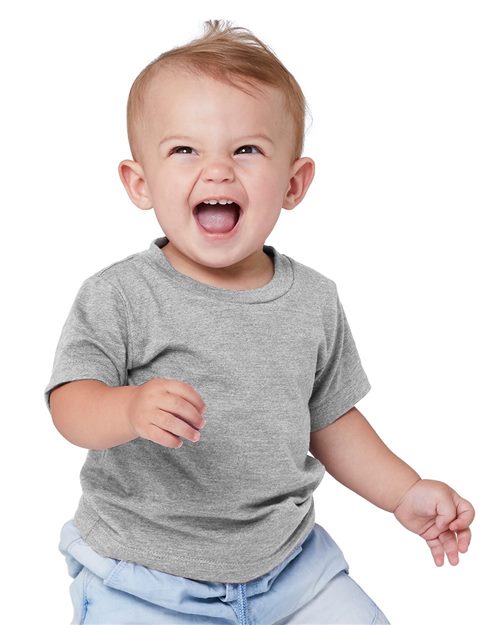 A joyful baby in a BELLA + CANVAS Infant Jersey Tee, made from Airlume combed and ring-spun cotton, sits against a white backdrop. With light-colored hair swept up and wearing light blue pants, the baby looks to the side with infectious charm.