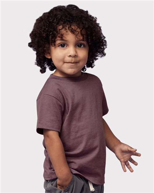 A young child with curly hair stands against a plain background, wearing a maroon BELLA + CANVAS - Toddler Jersey Tee and gray pants made from Airlume combed cotton, their left hand in their pocket and a slight smile on their face.