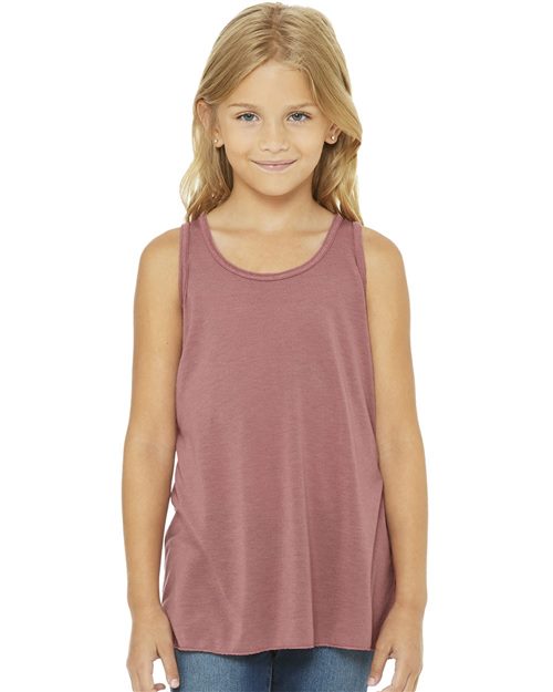 A young girl with long blonde hair is wearing a BELLA + CANVAS Youth Flowy Racerback Tank, made sustainably, and blue jeans. She stands smiling at the camera against a plain white background.