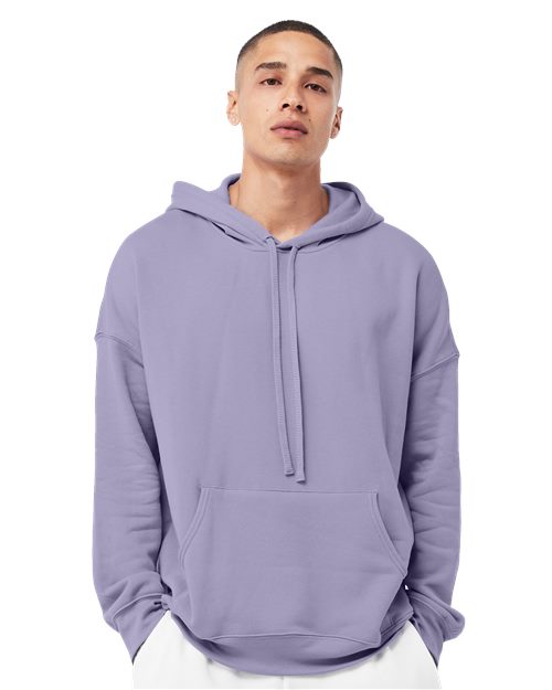 A person in a light purple BELLA + CANVAS Sponge Fleece Drop Shoulder Hoodie, crafted from Airlume combed and ring-spun cotton, stands against a white background. With unisex sizing, front pocket, drawstrings, and sustainable manufacturing highlighted by their neutral expression.