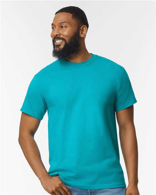 A bearded person smiles while looking sideways, wearing a bright turquoise Gildan Hammer™ T-Shirt made from OEKO-TEX certified ring-spun cotton and jeans against a plain white background.