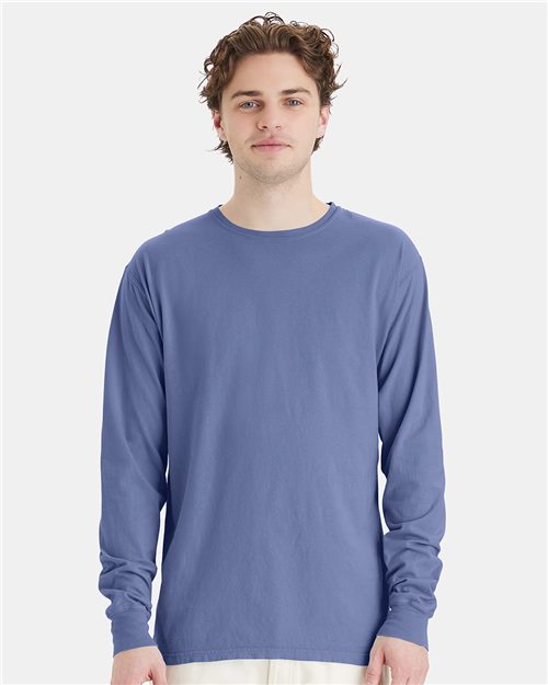 Load image into Gallery viewer, Against a light grey background, a person with wavy brown hair faces forward with a neutral expression, wearing white pants and a ComfortWash by Hanes Garment-Dyed Long Sleeve T-Shirt in blue, crafted through socially conscious manufacturing.
