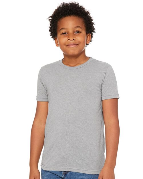 A curly-haired child smiles in a BELLA + CANVAS Youth Triblend Tee, made with Blue Sign certified dyes, and blue jeans, standing against a white backdrop.