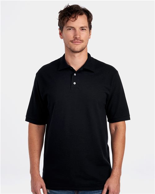 Someone with short, wavy hair wears a JERZEES - 100% Ring-Spun Cotton Piqué Polo in black. Its pre-shrunk fabric ensures a snug fit, featuring a collar and partially undone buttons. A plain background accentuates their neutral expression as they face the camera.