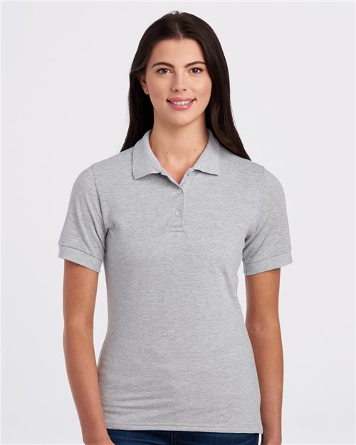 A person with long dark hair wears a JERZEES Women's 100% Ring-Spun Cotton Piqué Polo in light gray and jeans, standing against a plain white background. They smile at the camera, exemplifying sustainable manufacturing.