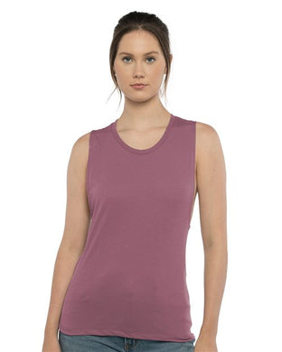 A person wears a Next Level - Women’s Festival Muscle Tank in mauve paired with blue jeans, standing against a plain white background. Their hair is pulled back as they look directly at the camera, embodying socially conscious manufacturing through every detail.