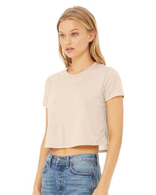 A woman with long blonde hair is wearing a light beige BELLA + CANVAS Women’s Flowy Crop Tee and blue jeans, standing against a plain white background.