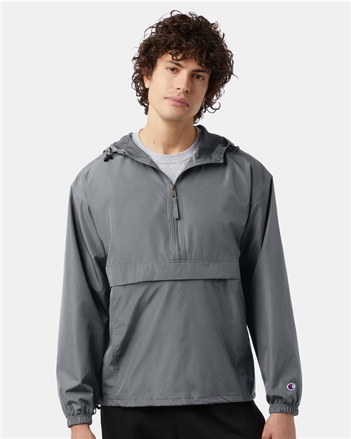 A person with curly hair is wearing a Champion Packable Anorak Jacket, a gray hooded windbreaker featuring an adjustable waist drawcord and wind- and water-resistant fabric. A small sleeve logo adorns it as they stand against a plain white background, embodying Sustainable Style.