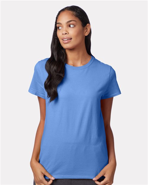 A person with long, dark hair wears a plain Hanes Perfect-T Women’s T-Shirt in blue, made from socially conscious manufacturing. Standing against a light gray background, they look to the side with hands at their waist.